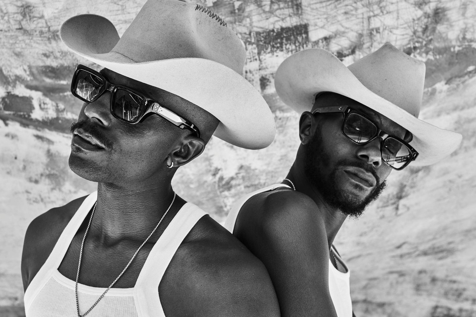 Two men in white cowboy hats, sunglasses and white tank tops pose back to back in black and white