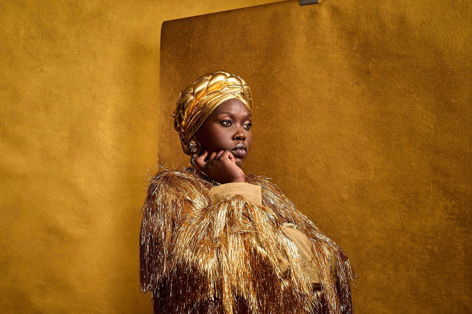 Woman in a gold braided headwrap and gold fringe jacket, chin resting on her hand against a gold backdrop