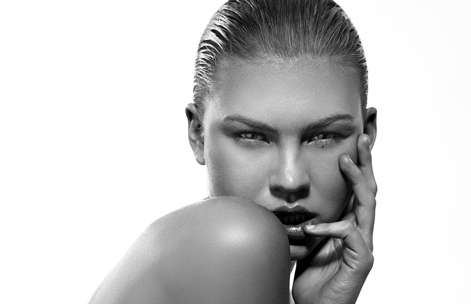 Black and white beauty portrait of a woman with slicked-back hair, fingers at her cheek, chin resting on her shoulder