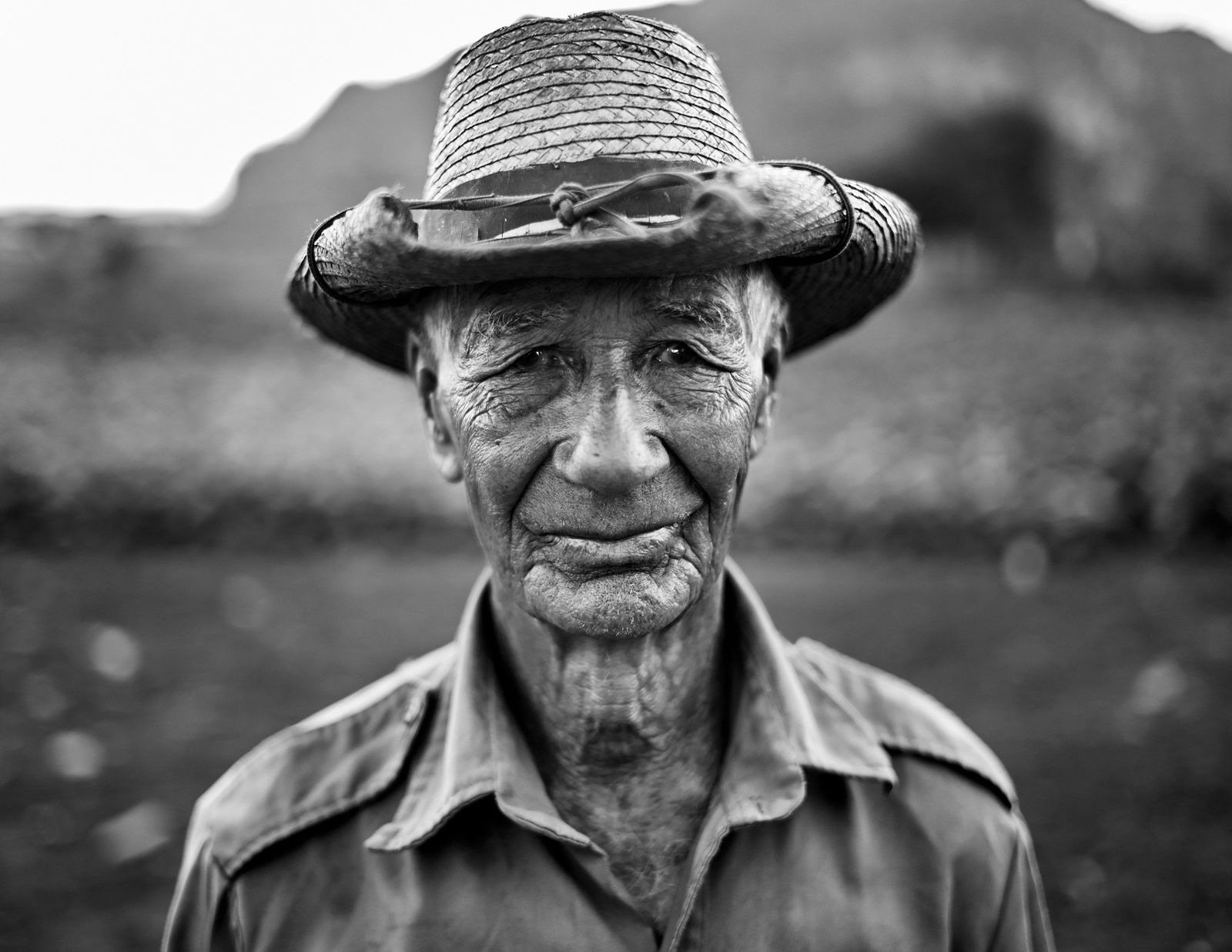 Black and white portrait of an older man in a worn straw hat, his face deeply lined, fields blurred behind